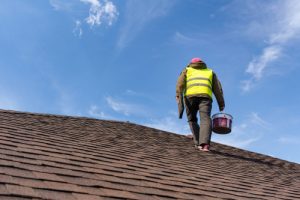 A roofer standing on top of a roof holding a bucket of roofing materials